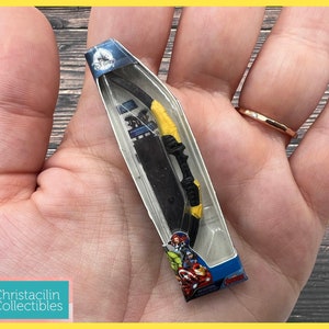 May include: A miniature toy bow and arrow set in a clear plastic package. The bow is black with yellow accents, and the arrow is black. The packaging features the Avengers logo and Disney branding.