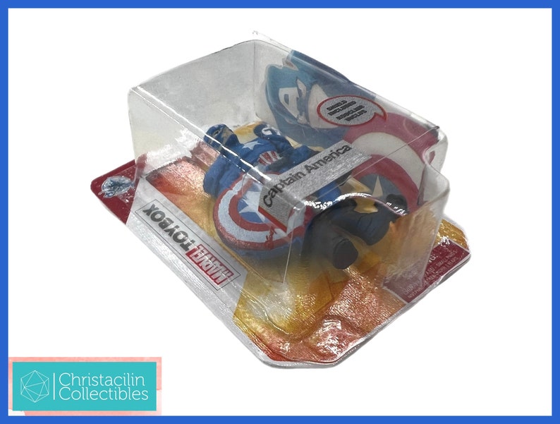 Op de afbeelding: Een Captain America actiefiguur in een doorzichtige plastic verpakking. Het figuur zit met een schild. De verpakking bevat de tekst "Captain America" en "Marvel Toybox". Het schild is rood, wit en blauw.