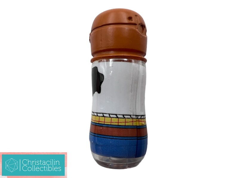 May include: A clear glass salt or pepper shaker with a brown top. The body of the shaker is white with a black cowhide pattern and a blue, yellow, and red design. The shaker is a collectible item.