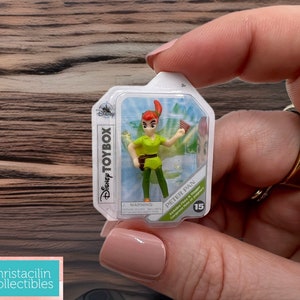May include: A Disney Toybox Peter Pan figure in a clear plastic package. The figure is wearing a green tunic, brown belt, and green pants. The packaging includes the Disney Toybox logo and the Peter Pan name.