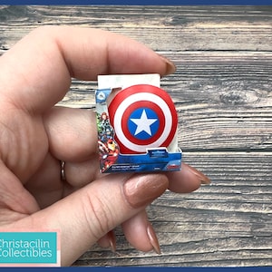 May include: A miniature Captain America shield, red with white stripes and a blue star, is displayed in its original packaging. The packaging features the Avengers and the words "Captain America's Shield". The shield is held in a person's hand.