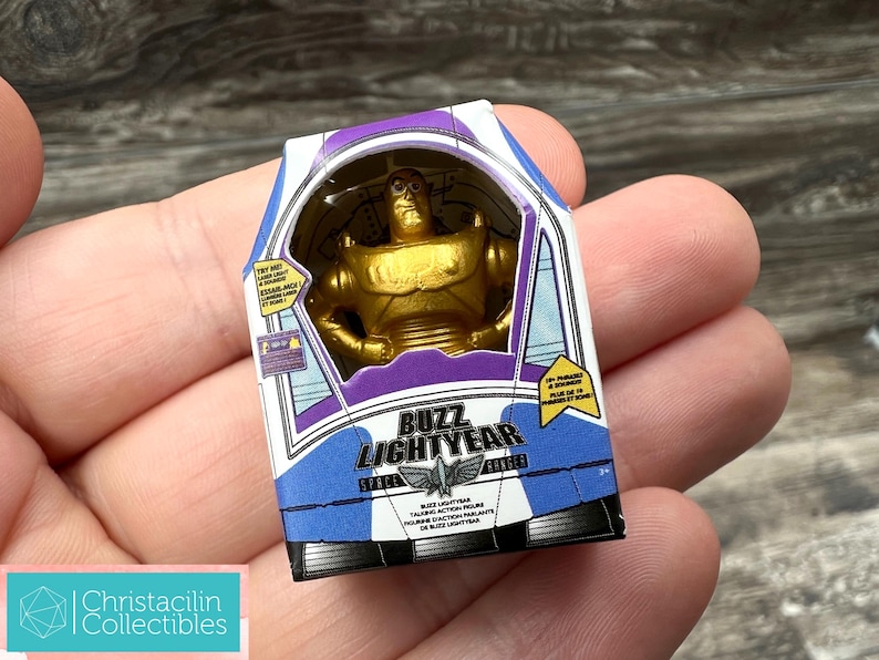 Puede incluir: Una figura de acci&oacute;n de Buzz Lightyear dorada en una caja. La caja es blanca, morada y azul con las palabras "Buzz Lightyear" y un gr&aacute;fico de una nave espacial. La caja tambi&eacute;n tiene texto en franc&eacute;s.
