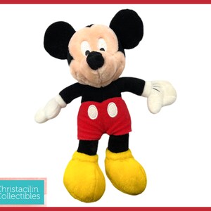 May include: A plush Mickey Mouse toy. Mickey has black ears, a tan face, black arms, white gloves, red shorts with two white buttons, and yellow shoes. The toy is set against a white background with a red border.