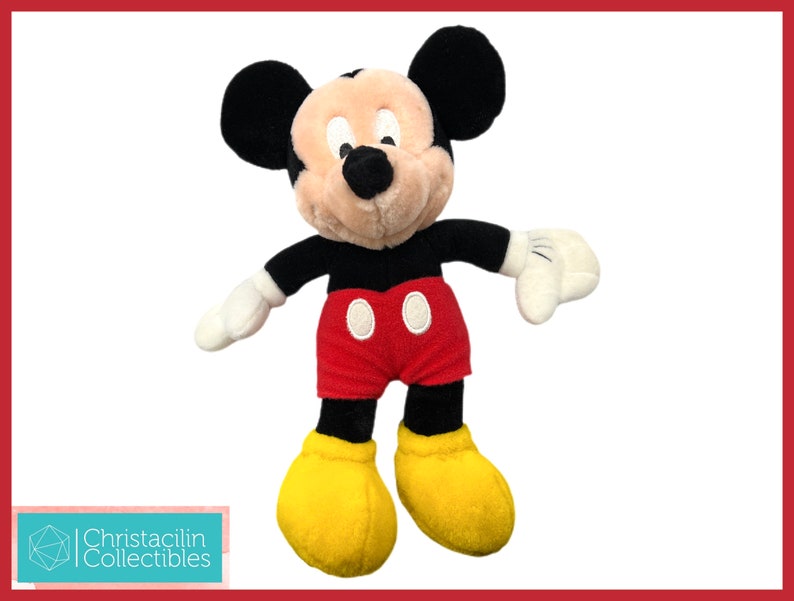 May include: A plush Mickey Mouse toy. Mickey has black ears, a tan face, black arms, white gloves, red shorts with two white buttons, and yellow shoes. The toy is set against a white background with a red border.
