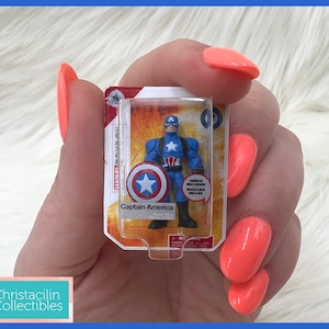 Op de afbeelding: Een miniatuur Captain America actiefiguur in een doorzichtige plastic verpakking. De figuur is blauw, rood en wit en houdt een schild vast. De verpakking leest "Captain America" en "Shield Included".