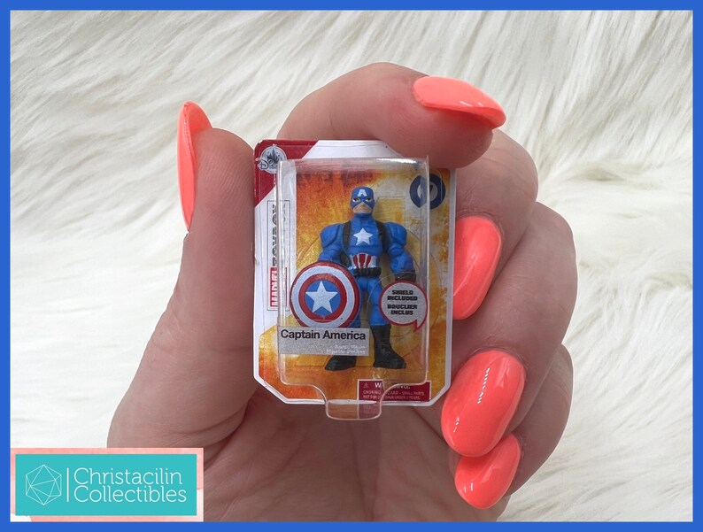 Op de afbeelding: Een miniatuur Captain America actiefiguur in een doorzichtige plastic verpakking. De figuur is blauw, rood en wit en houdt een schild vast. De verpakking leest "Captain America" en "Shield Included".