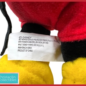 May include: A plush toy featuring red, black, and yellow colors. The toy has a tag with the Disney logo and text that reads "ALL NEW MATERIALS: POLYESTER FIBERS, PLASTIC PELLETS, EVA FOAM". The tag also includes the Walt Disney Parks and Resorts address.