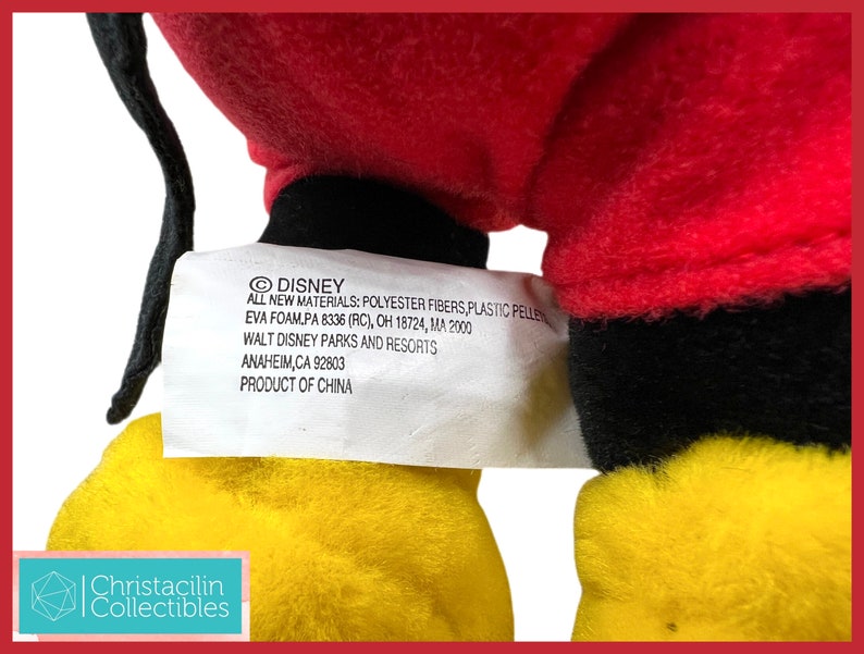 May include: A plush toy featuring red, black, and yellow colors. The toy has a tag with the Disney logo and text that reads "ALL NEW MATERIALS: POLYESTER FIBERS, PLASTIC PELLETS, EVA FOAM". The tag also includes the Walt Disney Parks and Resorts address.