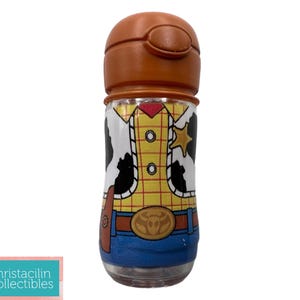 May include: A clear, cylindrical water bottle with a brown lid. The bottle is decorated with a design resembling the character Woody from Toy Story, featuring a yellow shirt, blue jeans, and a sheriff's badge.