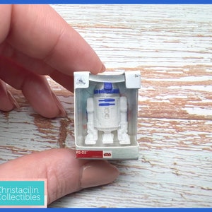 May include: A small, white and blue R2-D2 figurine in a clear plastic display case. The Star Wars collectible is in a rectangular box with the text "R2-D2" and the Star Wars logo. The figurine is held by a person.