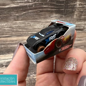 May include: A black toy race car with blue and white accents, labeled "XRS", sits in a small box. The box features a picture of a red and yellow race car. The toy car is held in a person's hand.