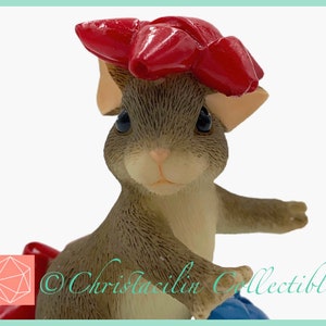 May include: A detailed figurine of a mouse wearing a large red bow on its head. The mouse has a light brown coat and is holding a blue object. The figurine is a collectible item.