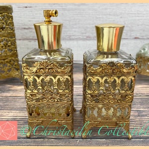 May include: Two antique perfume bottles with ornate gold filigree detailing. The bottles have clear glass bodies and gold-colored metal tops. The design includes intricate scrollwork and a rectangular shape. The bottles are displayed on a wooden surface.