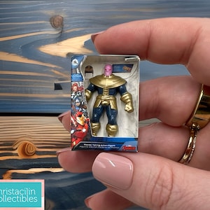 May include: Miniature Thanos action figure in its original packaging. The figure is in a standing pose, wearing a gold and blue costume. The packaging features Avengers characters. The figure is approximately 2 inches tall.