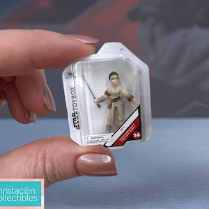 May include: A miniature Rey action figure from the Star Wars Toybox collection, held in a clear plastic package. The figure is dressed in tan and brown attire and holds a blue lightsaber. The package has the Star Wars Toybox logo.