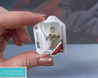 May include: A miniature Rey action figure from the Star Wars Toybox collection, held in a clear plastic package. The figure is dressed in tan and brown attire and holds a blue lightsaber. The package has the Star Wars Toybox logo.