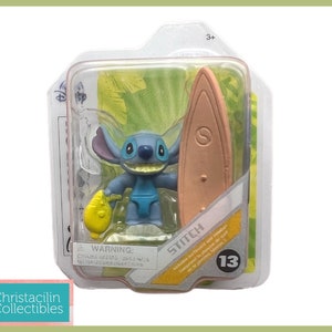 May include: A blue Stitch figurine with a surfboard and a yellow object, packaged in a clear plastic case. The packaging includes the Disney logo and the word "Stitch". The figurine is part of a collectible series.