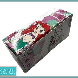 Op de afbeelding: Een rechthoekige doos met het Disney-personage Ariel uit De Kleine Zeemeermin. De doos heeft een doorzichtige plastic bovenkant en is versierd met een kleurrijk patroon van roze, groen en paars. De doos heeft ook de tekst "Christacilin Collectibles".