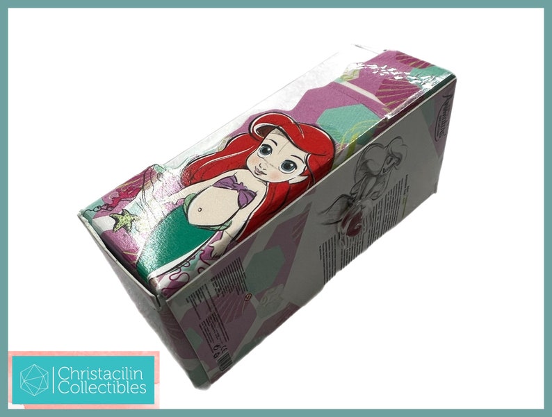 Op de afbeelding: Een rechthoekige doos met het Disney-personage Ariel uit De Kleine Zeemeermin. De doos heeft een doorzichtige plastic bovenkant en is versierd met een kleurrijk patroon van roze, groen en paars. De doos heeft ook de tekst "Christacilin Collectibles".