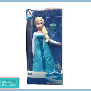 Pu&ograve; includere: Una statuetta di Elsa dal film Disney Frozen, confezionata in una scatola di plastica trasparente. La figura indossa un abito azzurro con un top bianco e una treccia bionda. La scatola presenta il logo Disney Frozen.