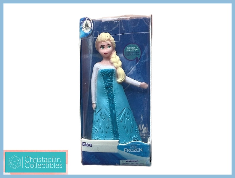 Pu&ograve; includere: Una statuetta di Elsa dal film Disney Frozen, confezionata in una scatola di plastica trasparente. La figura indossa un abito azzurro con un top bianco e una treccia bionda. La scatola presenta il logo Disney Frozen.