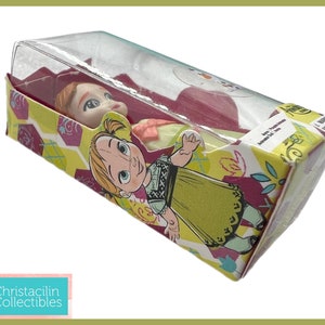 May include: A doll of the character Anna from the movie Frozen, packaged in a colorful box. The box features illustrations of Anna and floral patterns in yellow, purple, and green. The doll is visible through a clear plastic window.