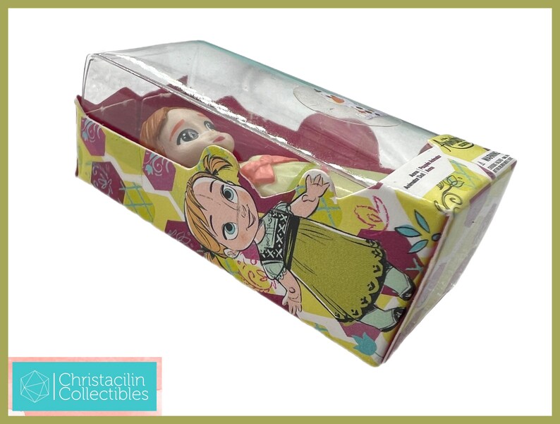 May include: A doll of the character Anna from the movie Frozen, packaged in a colorful box. The box features illustrations of Anna and floral patterns in yellow, purple, and green. The doll is visible through a clear plastic window.