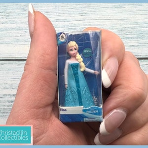 Pu&ograve; includere: Una bambola in miniatura di Elsa dal film Disney Frozen, confezionata in una scatola di plastica trasparente. La bambola indossa un vestito blu con un top bianco e ha i capelli biondi. La scatola presenta il logo Disney Frozen e il nome "Elsa".