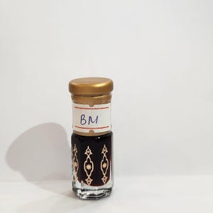 May include: A small glass bottle with a gold lid and a white label that says "BM". The bottle is filled with a dark liquid and has a decorative pattern on the side.