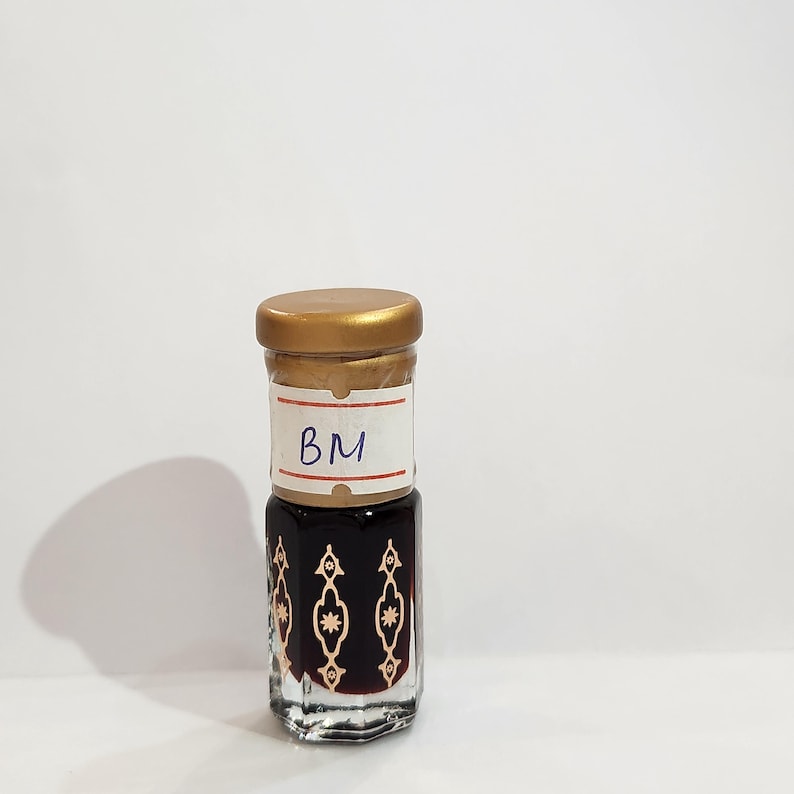 May include: A small glass bottle with a gold lid and a white label that says "BM". The bottle is filled with a dark liquid and has a decorative pattern on the side.