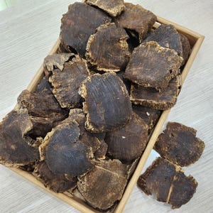 May include: A wooden tray filled with numerous dark brown, irregularly shaped pieces of wood. The wood pieces have a rough, textured surface and varying shades of brown. The tray is made of light-colored wood, and the background is a light wood surface.