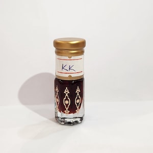 May include: A small, clear glass bottle with a gold lid and a white label that says "KK". The bottle is filled with a dark brown liquid and has a decorative pattern on the glass.