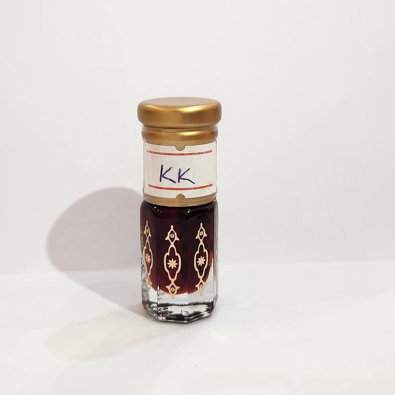 May include: A small, clear glass bottle with a gold lid and a white label that says "KK". The bottle is filled with a dark brown liquid and has a decorative pattern on the glass.