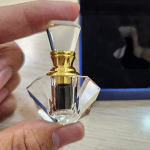 May include: A small, clear glass bottle with a gold cap and a diamond-shaped base. The bottle is filled with a dark liquid.