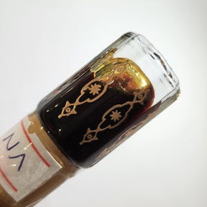 May include: A small glass bottle with a gold and black label. The bottle is filled with a dark liquid and has a gold cap. The label says "N/A".