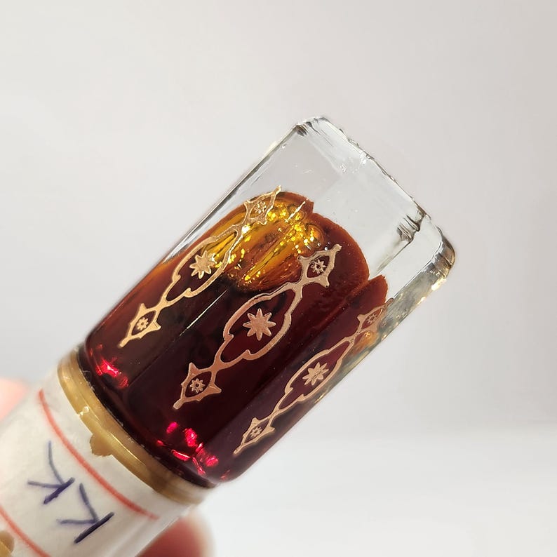 May include: A small glass bottle with a gold and red design. The bottle is filled with a dark red liquid. The bottle has a label with the letter "K" written on it.