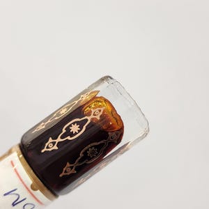 May include: A small glass bottle with a dark brown liquid inside. The bottle has a gold and black patterned design and a clear glass top.