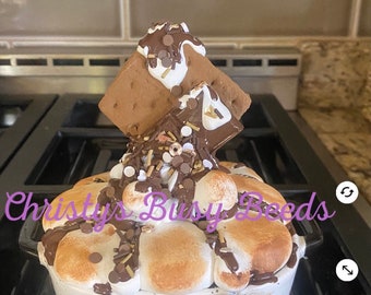 May include: A white ceramic bowl filled with toasted marshmallows, chocolate drizzle, and sprinkles, topped with a s'more. A black and white gingham ribbon is tied around the front. The text Christys Busy Beeds is overlaid on the image.