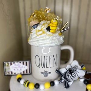 May include: White ceramic mug with the word "QUEEN" in black lettering and a small bee design. The mug is topped with whipped cream, honeycomb, and bee figurines. A black and white striped straw, a black and white bee-themed bow, and a yellow, black, and white beaded necklace are also pictured.