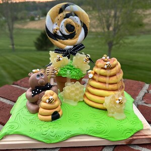 May include: A whimsical bee-themed tabletop decoration featuring a black and yellow lollipop, a brown bear figurine, and a honey comb structure. The scene is set on a green base with a wood platform.