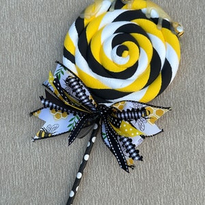 May include: A decorative lollipop ornament featuring a spiral of yellow, black, and white foam. The top is accented with two small bee figures and a translucent drip effect. A multi-layered ribbon bow with bee and honeycomb patterns sits below.