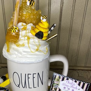 May include: White ceramic mug with the word "QUEEN" in black lettering. The mug is decorated with a beehive, honey, and bees. The mug is sitting on a white plate with yellow and white candy.