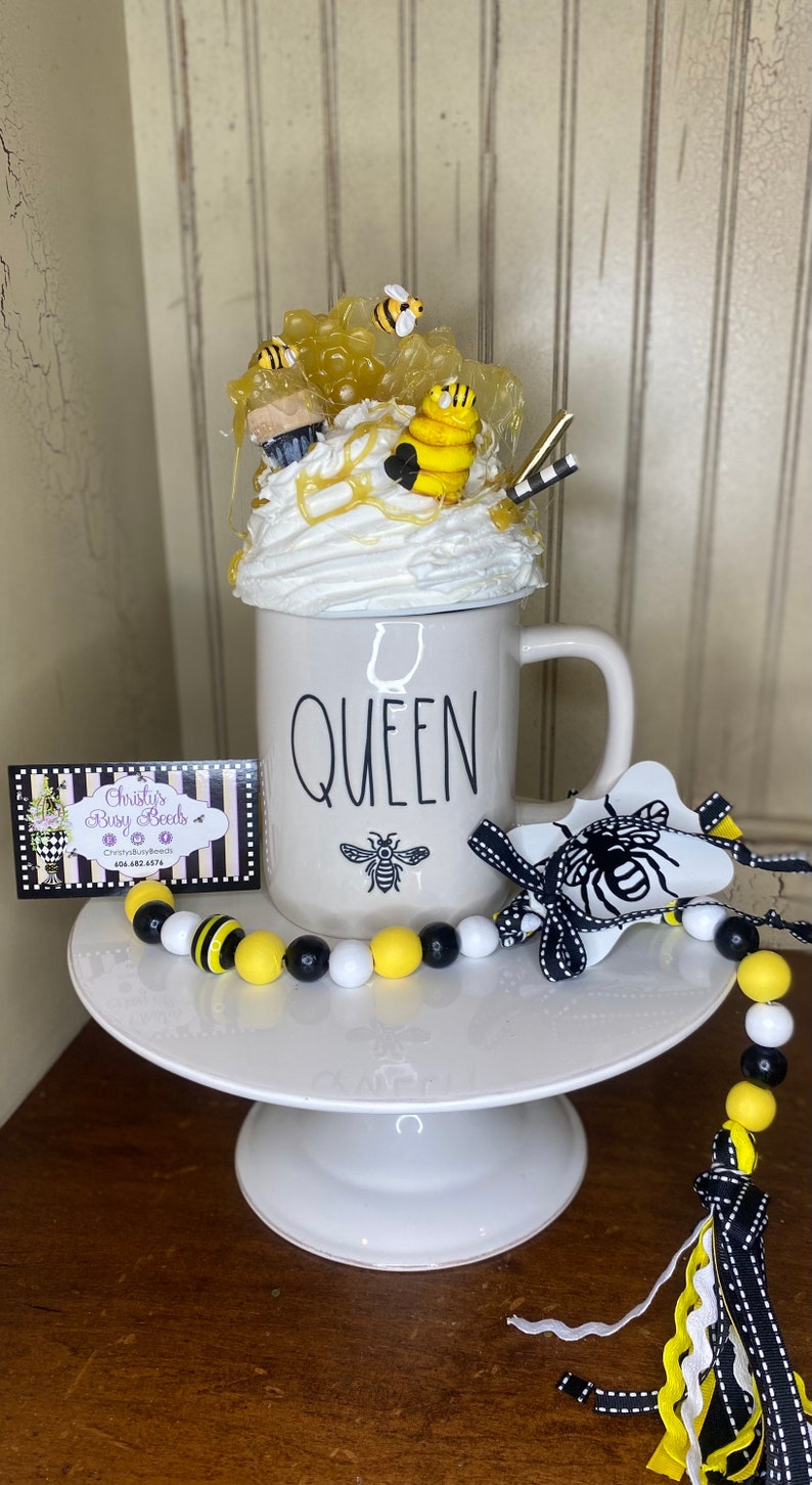 May include: White ceramic mug with the word "QUEEN" printed on it. The mug is decorated with a bee and honeycomb design. A black and yellow beaded garland with a black and white ribbon tassel is in the foreground.