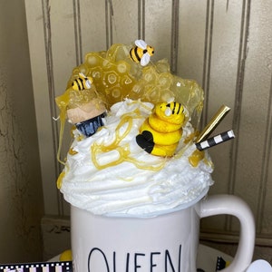May include: White ceramic mug with the word "QUEEN" in black lettering. The mug is decorated with a bee-themed design, including a yellow and black bee, honeycomb, and honey dripping down the side.
