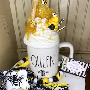 May include: A white mug with the word "QUEEN" in black lettering, decorated with bee-themed embellishments. The mug is topped with a whipped cream design, honey, and bee figurines. A decorative bee-themed garland and business card are also present.