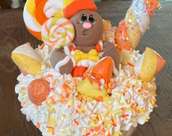 Puede incluir: Decoración en forma de calabaza con un hombre de jengibre rodeado de caramelos. La figura lleva un sombrero y una pajarita de maíz dulce, todo sobre glaseado blanco. Adorno con maíz dulce, piruletas y chispas, naranja, amarillo y blanco.