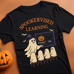 Puede incluir: Camiseta negra con el texto "SPOOKERVISED LEARNING" en naranja. La camiseta presenta un profesor fantasma de dibujos animados con un sombrero de bruja, enseñando a otros fantasmas sobre calabazas y murciélagos. Dos calabazas talladas están a la izquierda.