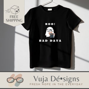 Può includere: Maglietta nera con una grafica di un fantasma con occhiali e libri. Il testo "BOO! BAD DATA" è sopra e sotto il fantasma. La maglietta è appesa a una gruccia bianca contro un muro bianco.