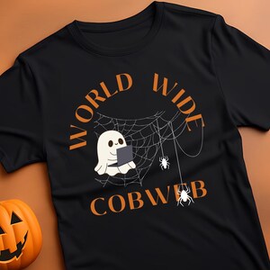 Op de afbeelding: Zwart T-shirt met een Halloween-ontwerp. Het shirt heeft een spook in een web met de woorden "WORLD WIDE COBWEB" in oranje. Een uitgesneden pompoen en kleine lantaarns staan links.