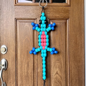 May include: A beaded lizard ornament in turquoise, blue, and pink hangs from a silver ring. The lizard is made of colorful beads and is suspended against a wooden door. The lizard's body is turquoise, with pink and blue accents.